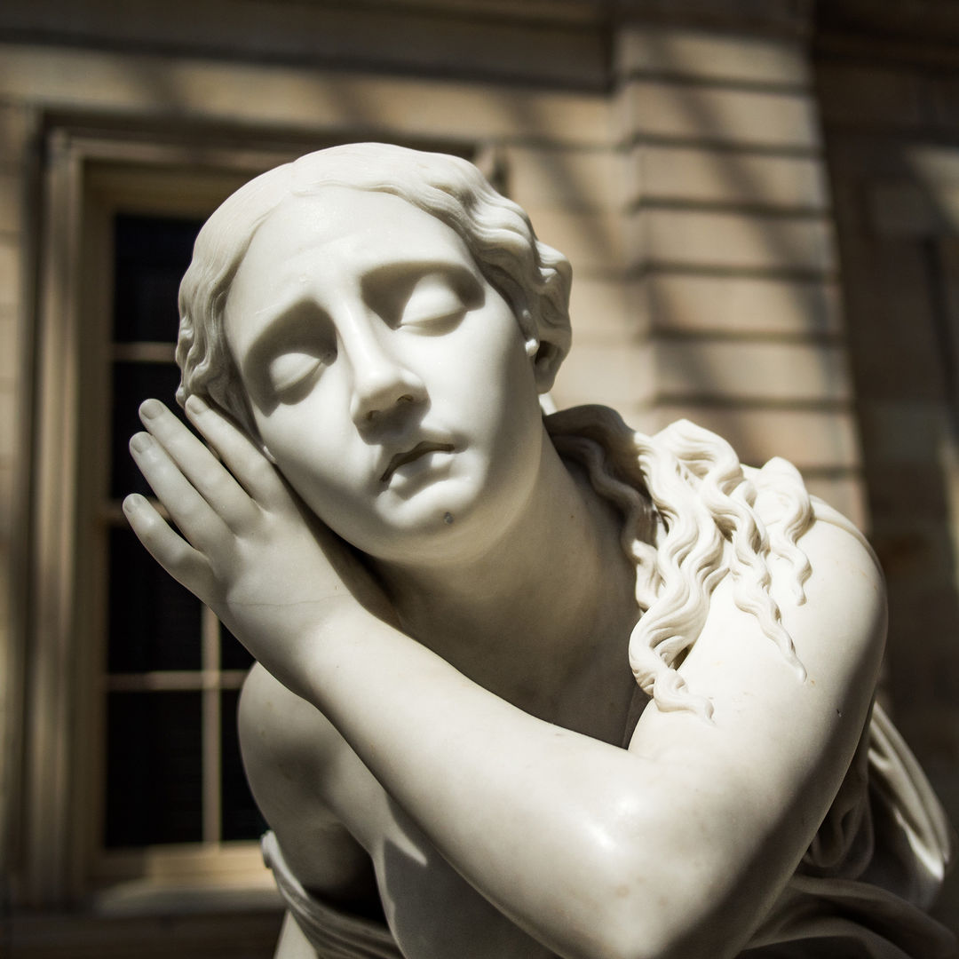 Close-up of the marble statue of Nydia, The Blind Flower Girl of Pompeii, made by Randolph Rogers, from her above her shoulders showing a young girl with closed eyes and a hand cupped around her right ear in a gesture suggesting it aids her hearing. Nydia’s face is directly facing the camera. The sculpture is in the American Wing Engelhard Sculpture Court at The Met, a skylit space with direct, dramatic natural light.