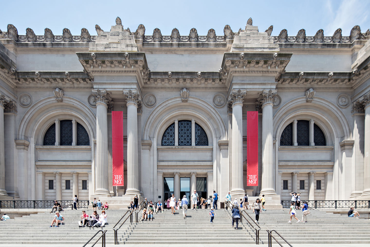 View of The Met facade, looking up from the exterior steps at 5th Ave. The building's columns, arched windows, and architectural treatment along the roofline are dramatic and imposing.