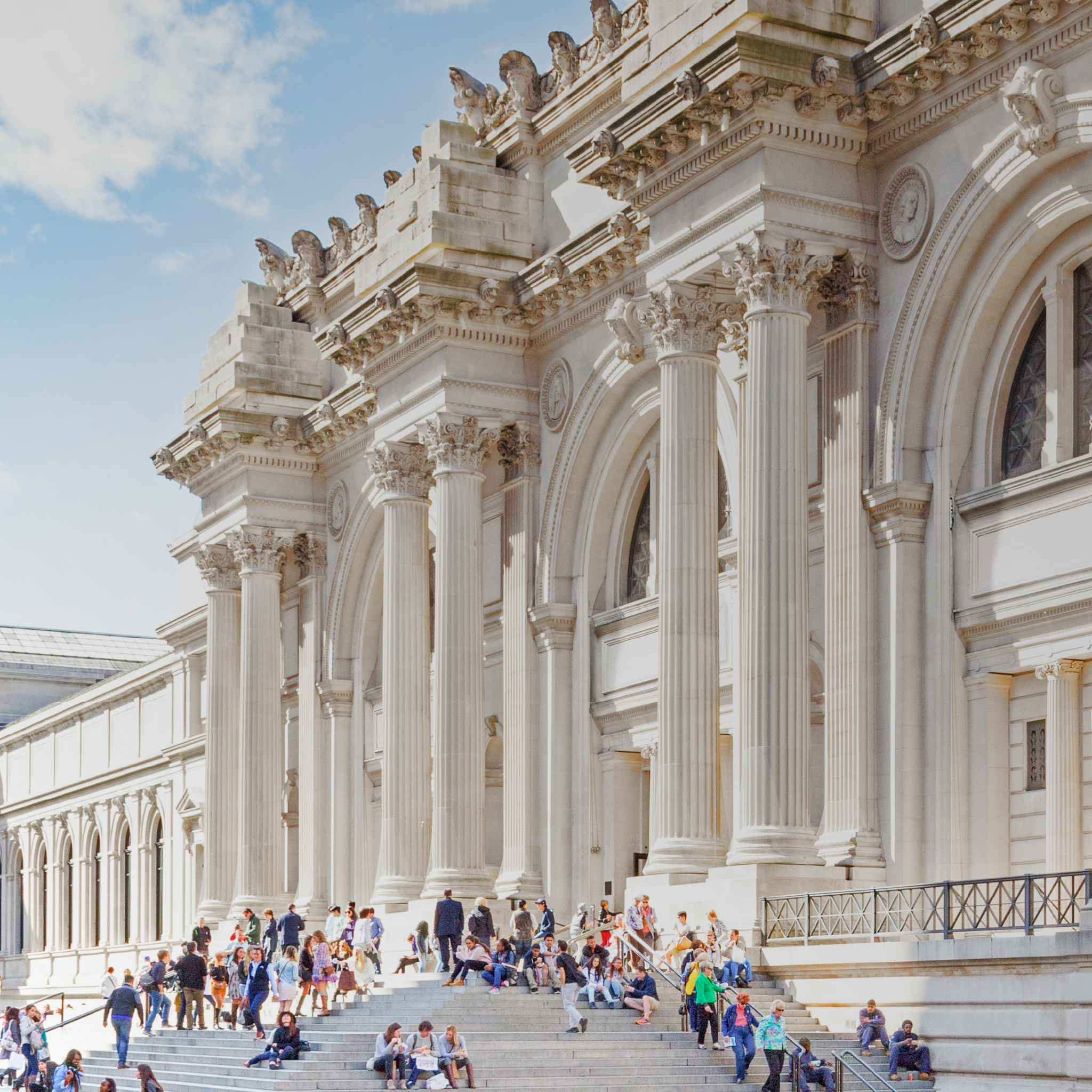 South-facing view of The Met exterior steps from the 5th Ave plaza. Sunlight hits the building facade, and the steps bustle with seated and standing visitors.