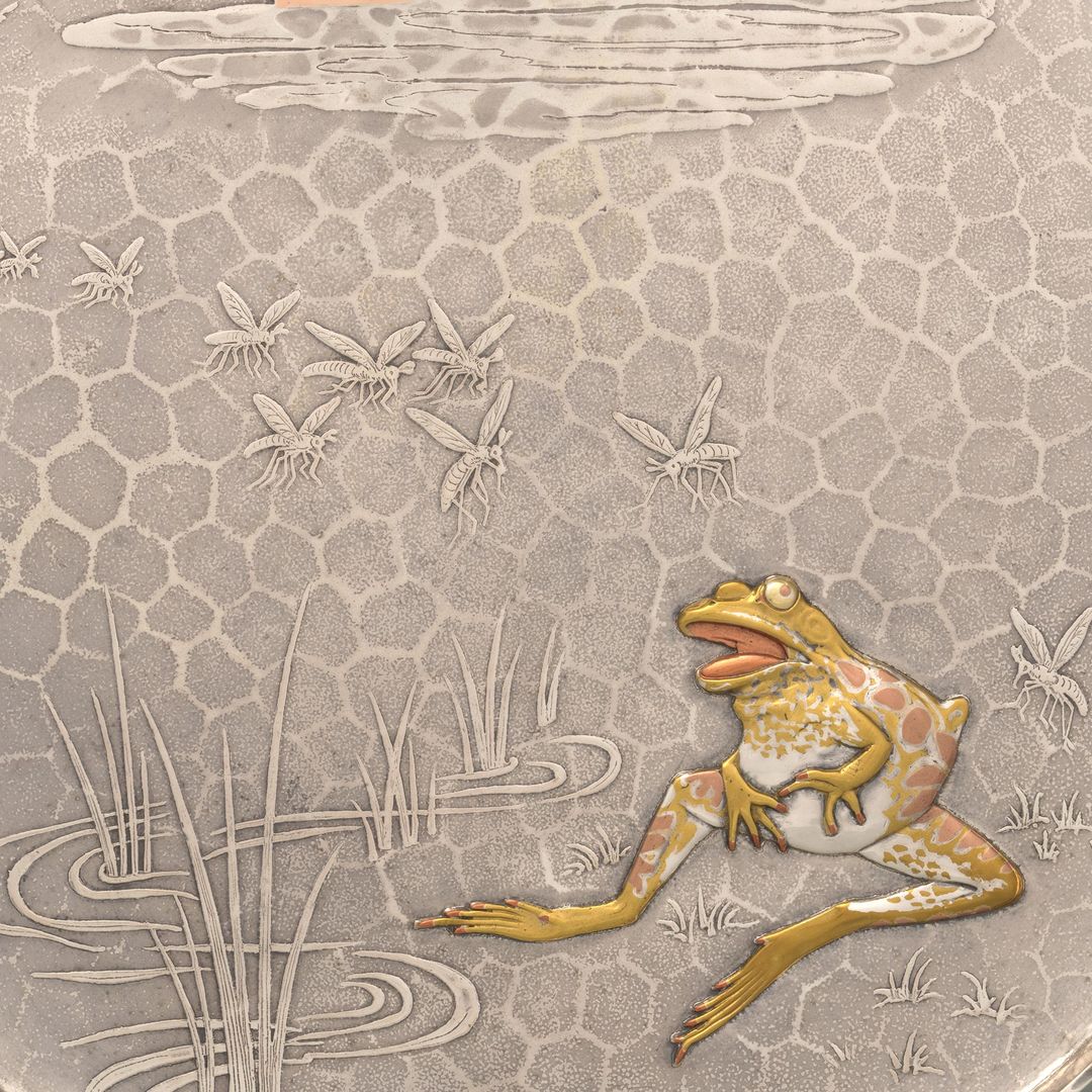 Detail of a silver tray featuring the design of a frog seated at the edge of a grassy pond with a queue of mosquitos approaching from a setting sun on the horizon. The surface has a hexagonal-shaped texture. The grass and mosquitoes protrude in a low relief on the tray surface. The front is more heavily sculpted and plated with mixed metals that are silver, gold, and copper in tone.