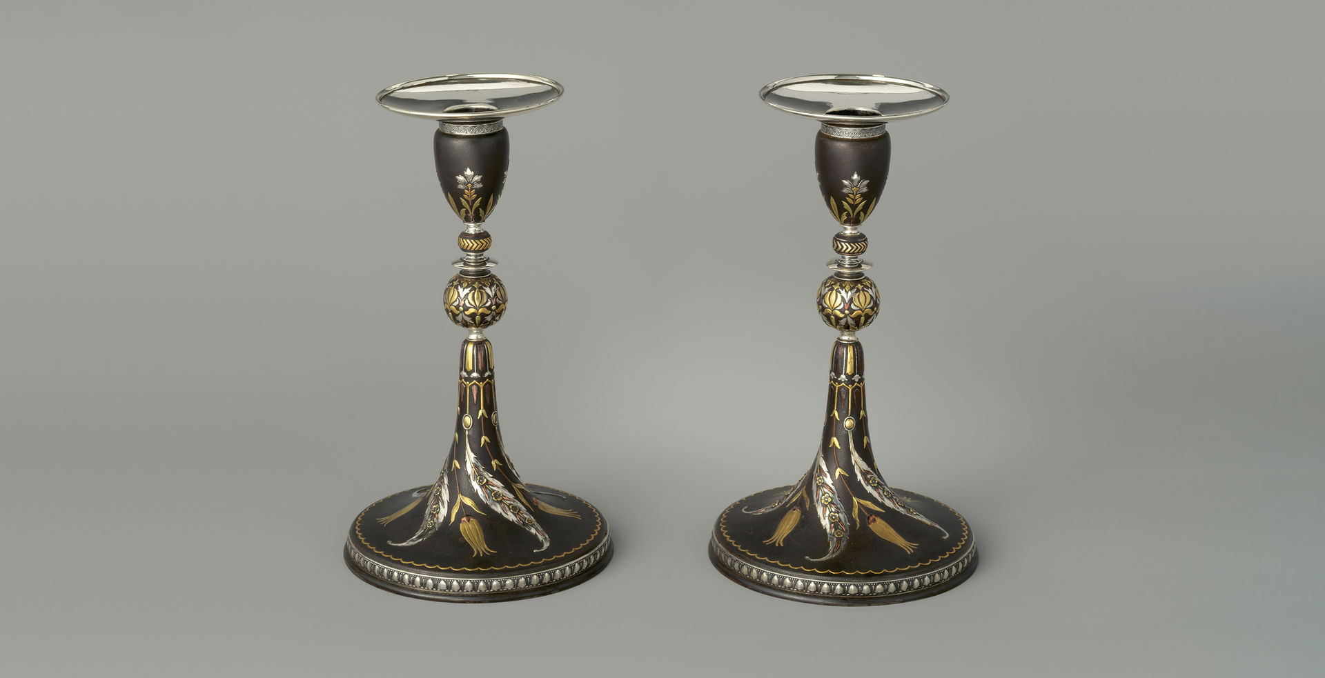 A pair of black iron candlesticks with a tapered conical base appear side-by-side. They are adorned with various sinuous floral motives that are modeled in low relief on the surface and surfaced in a variety of silver-, gold-, and copper-toned metals.