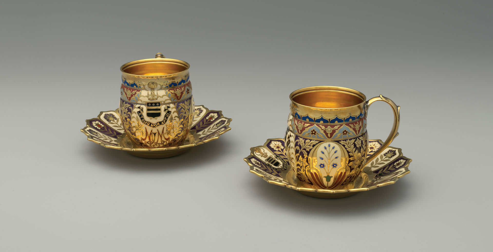 A pair of dazzling gilded and enameled cups and saucers adorned with floral designs, a coat of arms, and monogram.