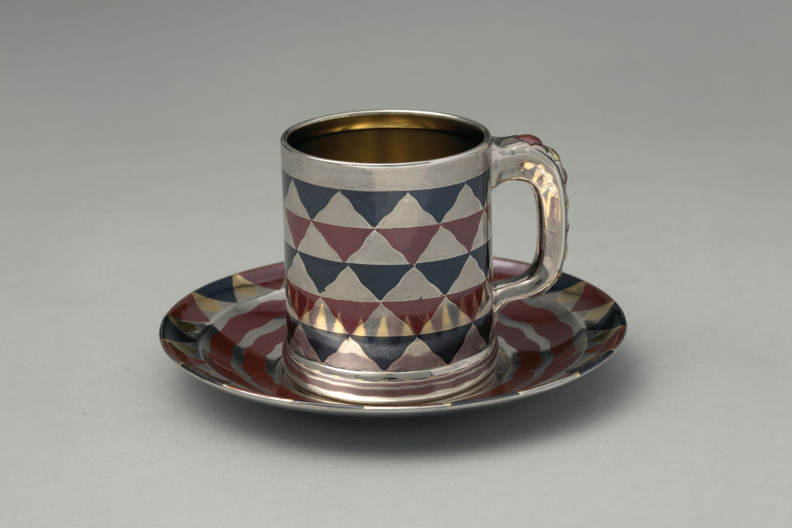 A silver, navy, and maroon cylindrical cup with a handle rests on a matching saucer. The cup and saucer are reflective and adorned with an inlay design of a checkerboard pattern of wavy triangle shapes.