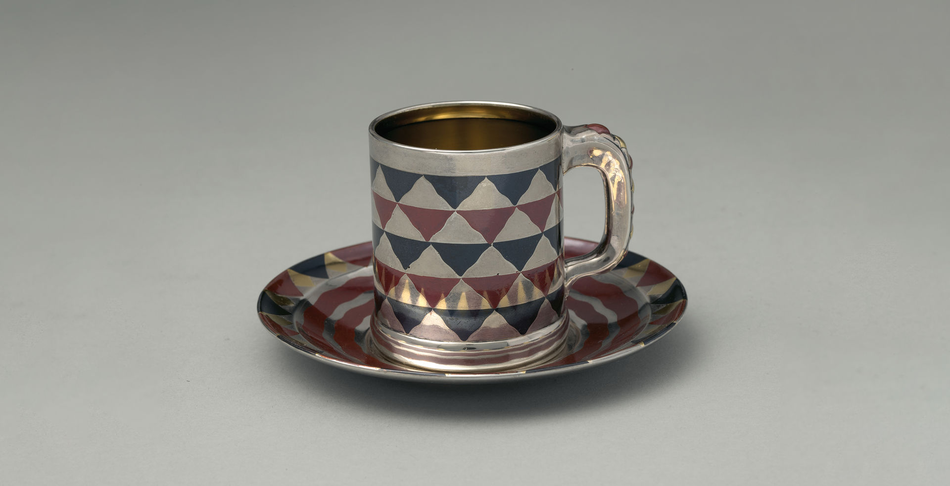 A silver, blue, and maroon cylindrical cup with a handle rests on a matching saucer. The cup and saucer are reflective and adorned with an inlay design of a checkerboard pattern of wavy triangle shapes.