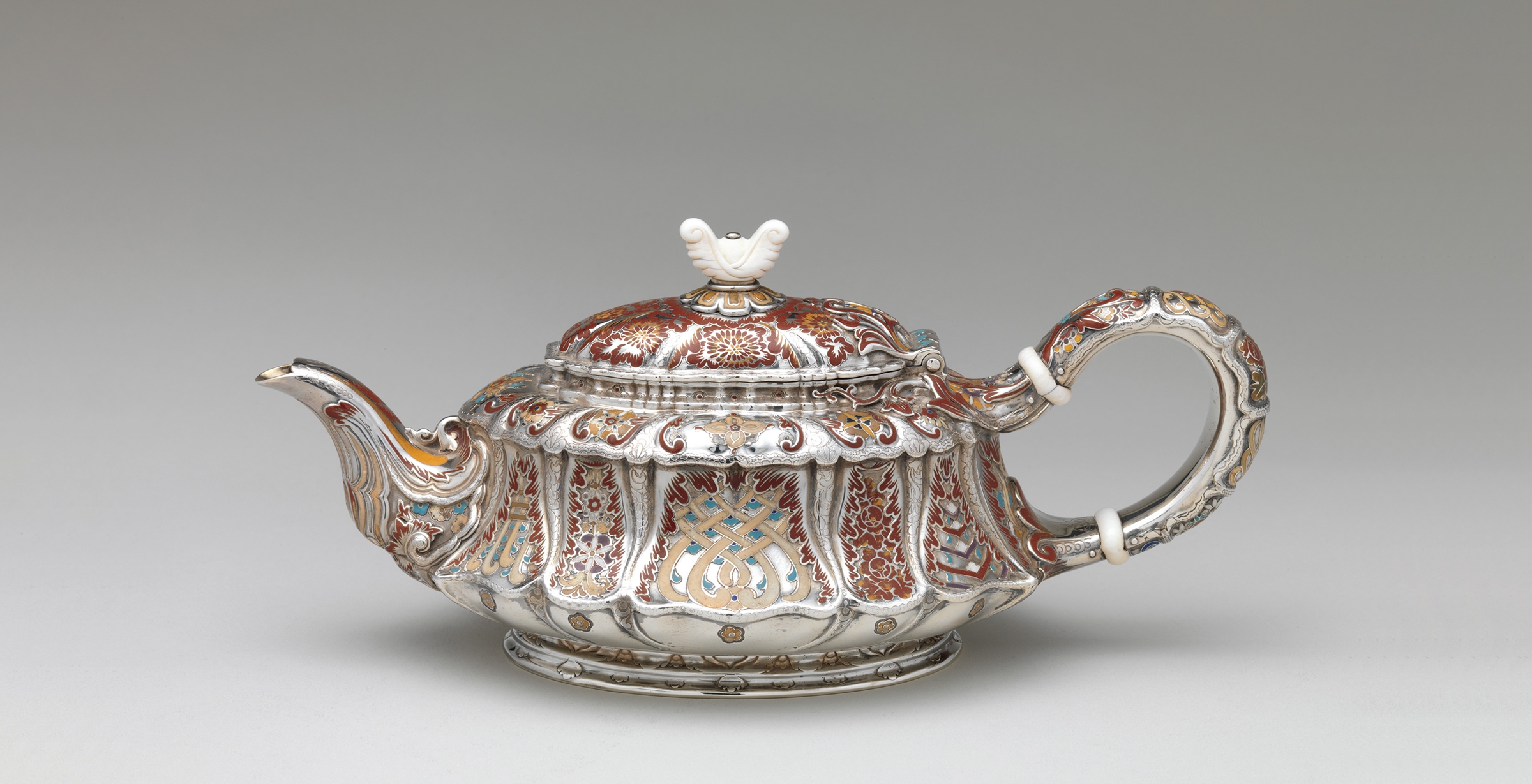 A short but elongated silver teapot with an ivory lid finial adorned with abstract floral motifs rendered in maroon, orange, taupe, and turquoise enamel.