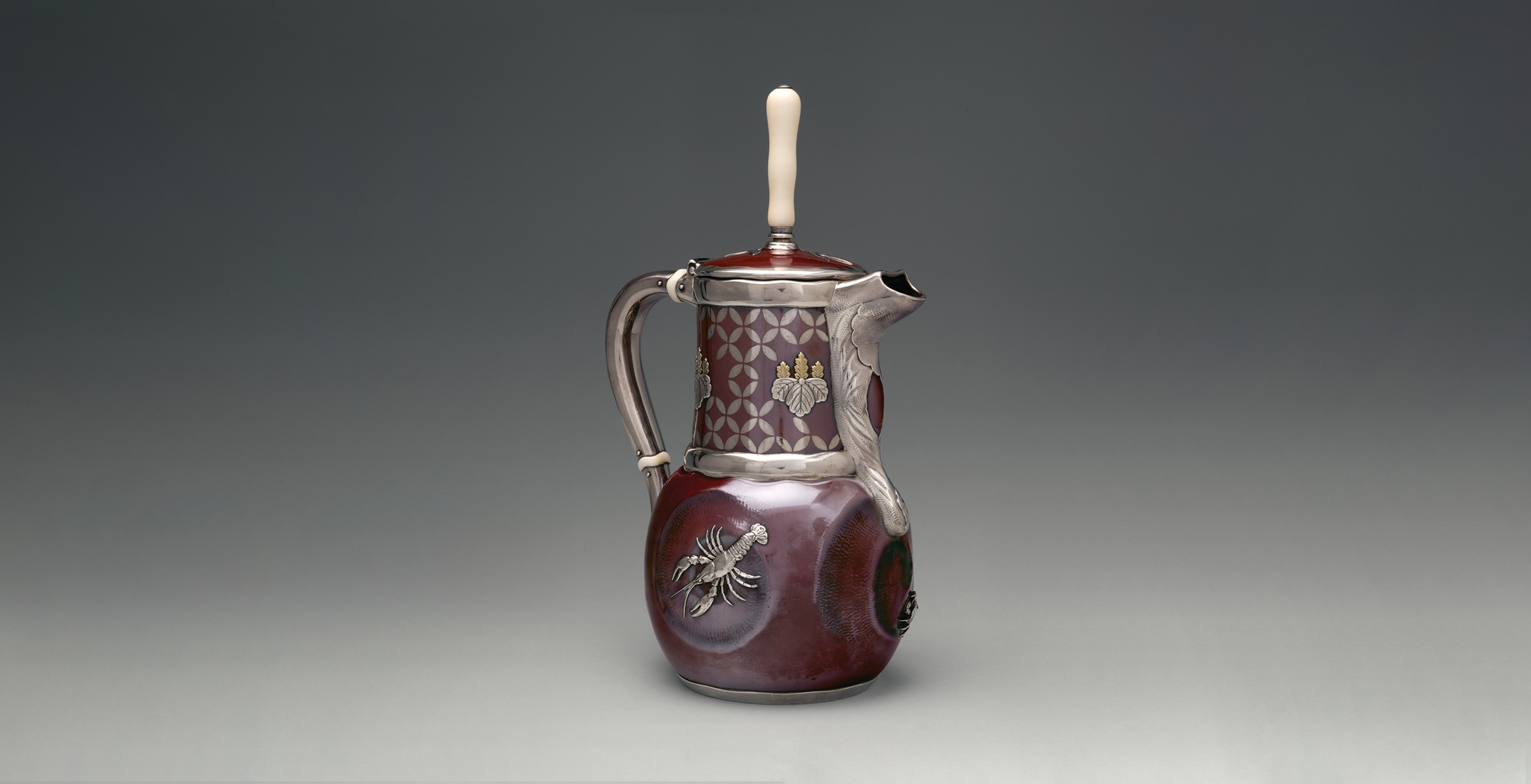 A three-quarter view of a patinated copper and silver chocolate pot in the shape of a pitcher with a handle, spot, and ivory handle. Silver banding emphasizes the handle, spout, and rim of the lid and neck. A lobster rendered in high-relief silver adorns the body of the pot.