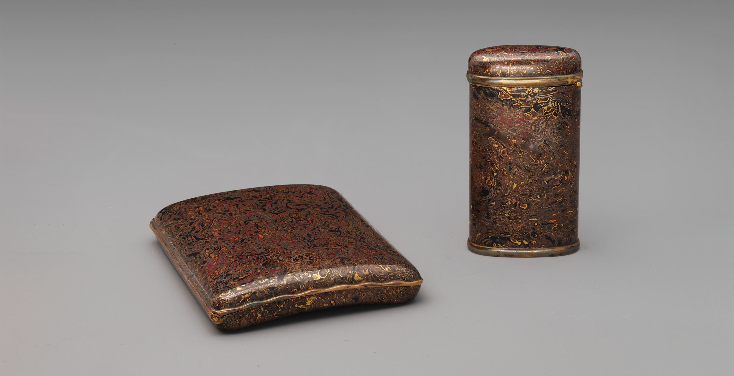 A flat cigarette case and a tall match case in a matching wood grain pattern rendered in deep red, black, brown, and gold metals. 