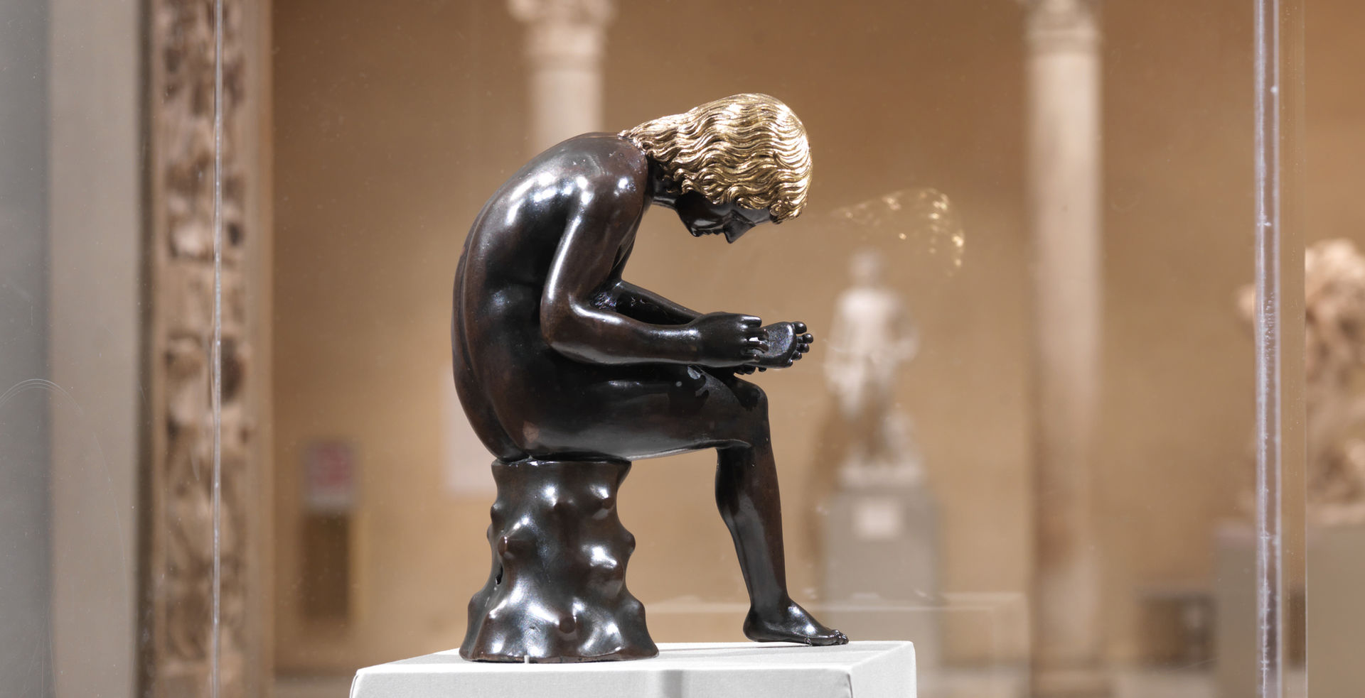 A small bronze sculpture of a seated boy pulling a thorn from the bottom of his foot appears within a display case at The Metropolitan Museum of Art; the figure hunches over looking intently at the bottom of his foot; the sculpture is set against the warm walls and limestone columns of the against gallery in the distance.