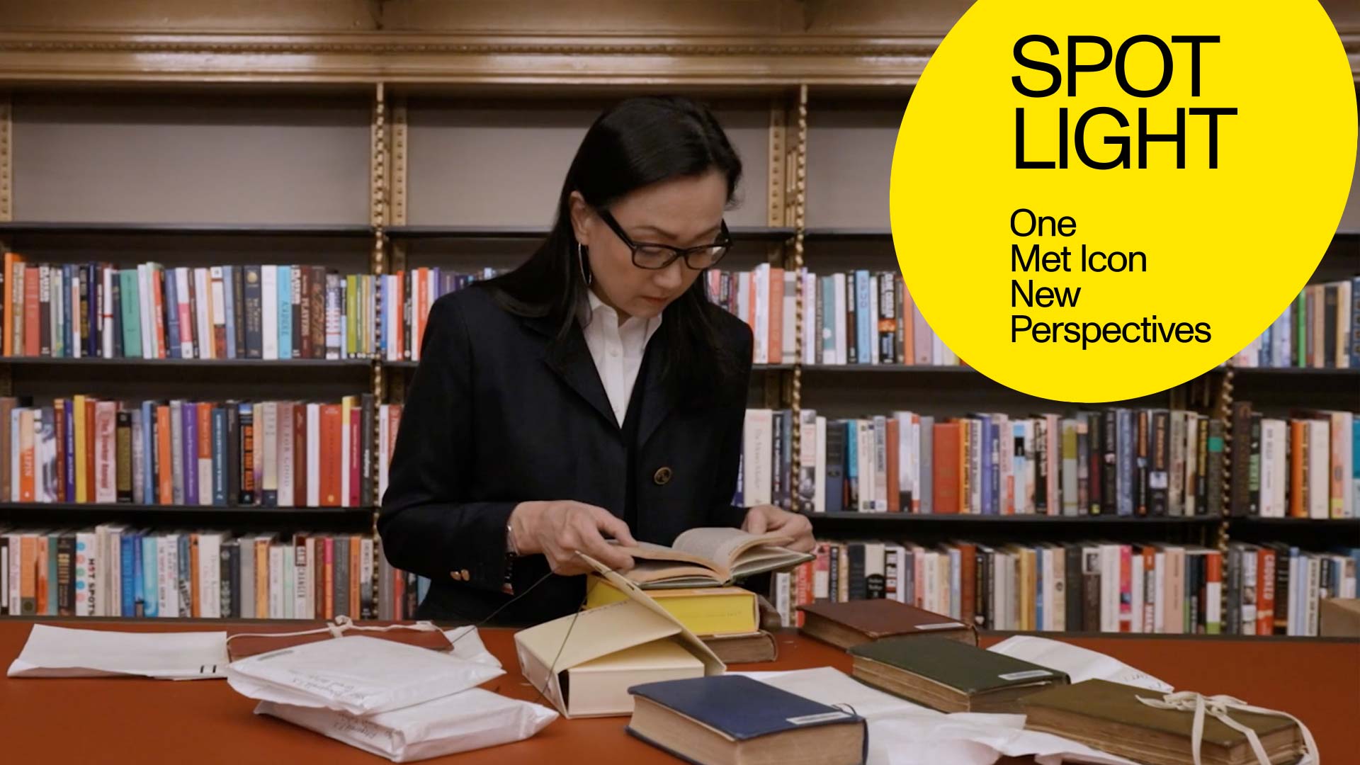 The Novelist Min Jin Lee peruses through an assortment of books atop a red-orange tabletop surface, set against a backdrop of book shelves in a New York Public Library study room.
