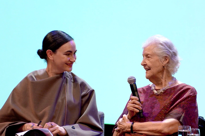 The architect Frida Escobedo smiling at artist Sheila Hicks, who holds a microphone. They are seated with a light blue background behind them.