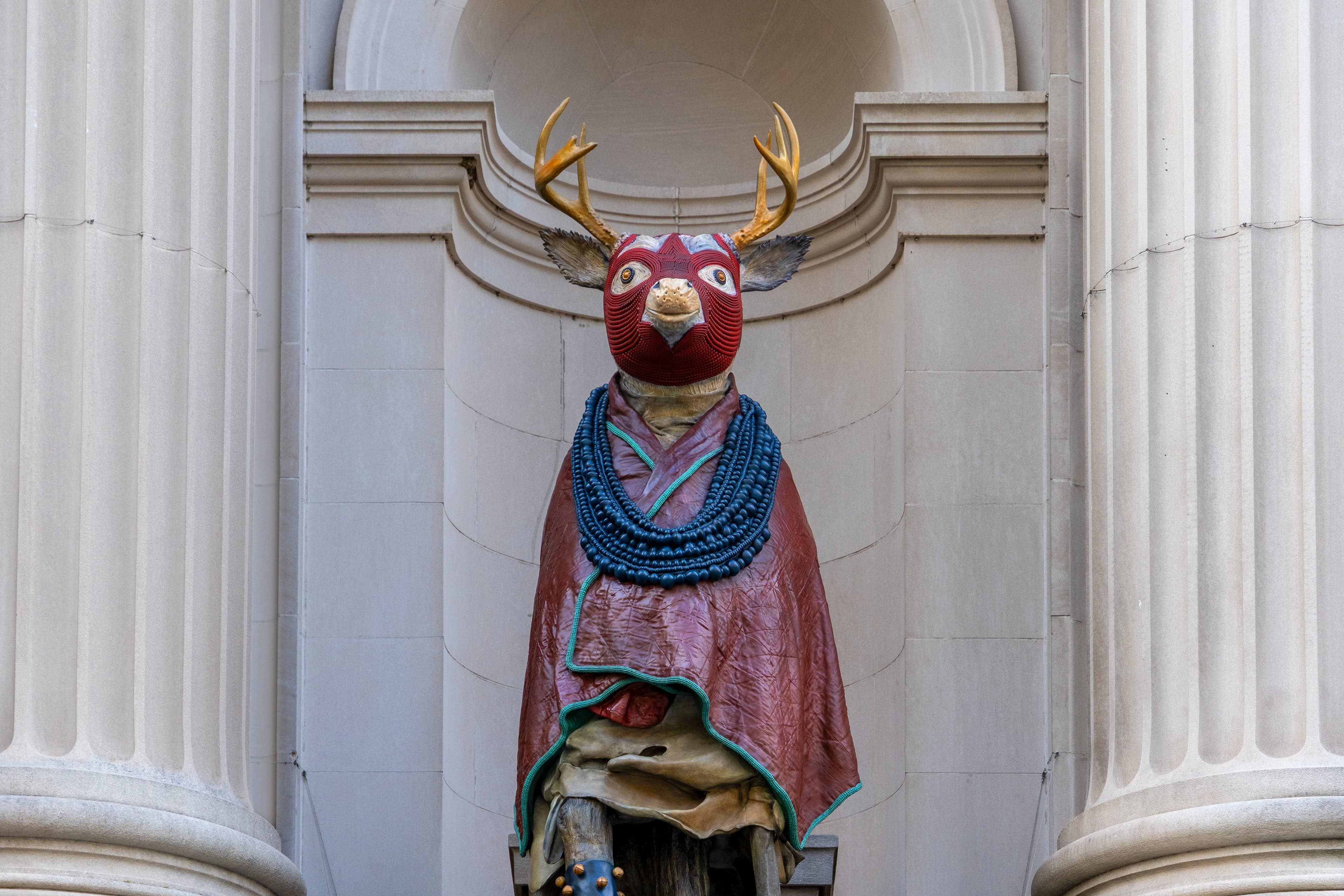 An image of a sculpture in the Met Facade. The sculpture is of a deer with a beaded mask and a cloak with a beaded necklace.