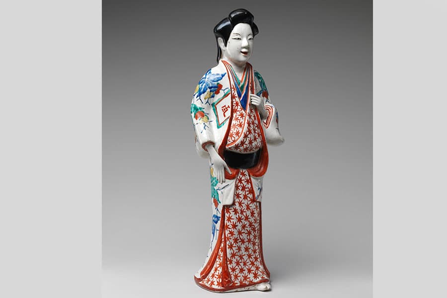 A Japanese female figure made of porcelain. She has black hair in an ornate hairstyle and is wearing a draping outfit that is largely white with red, blue, and green detailing.