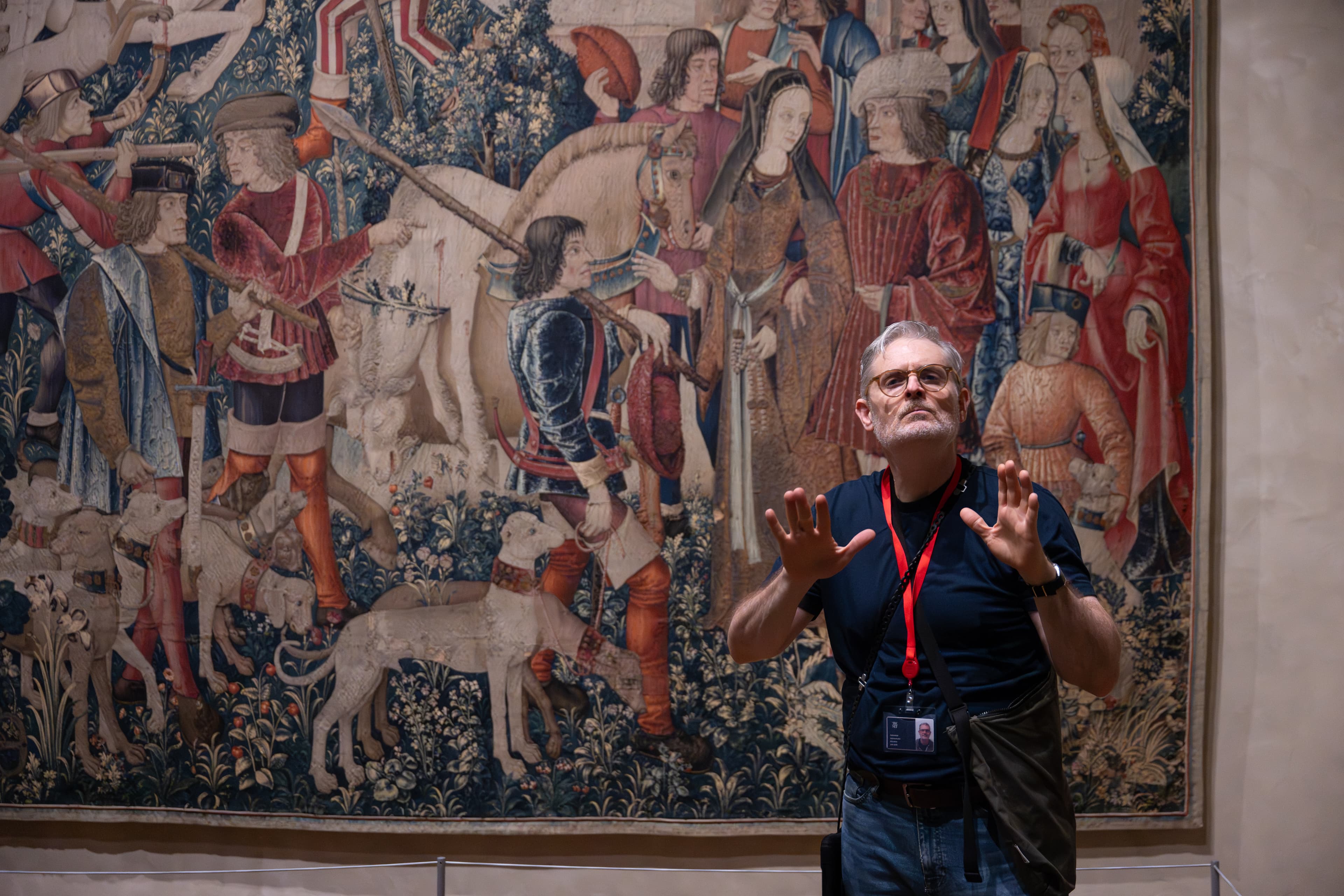 A Deaf educator stands in front of a Medieval tapestry with his hands up, at about chest height, as he signs. His face is quite serious.