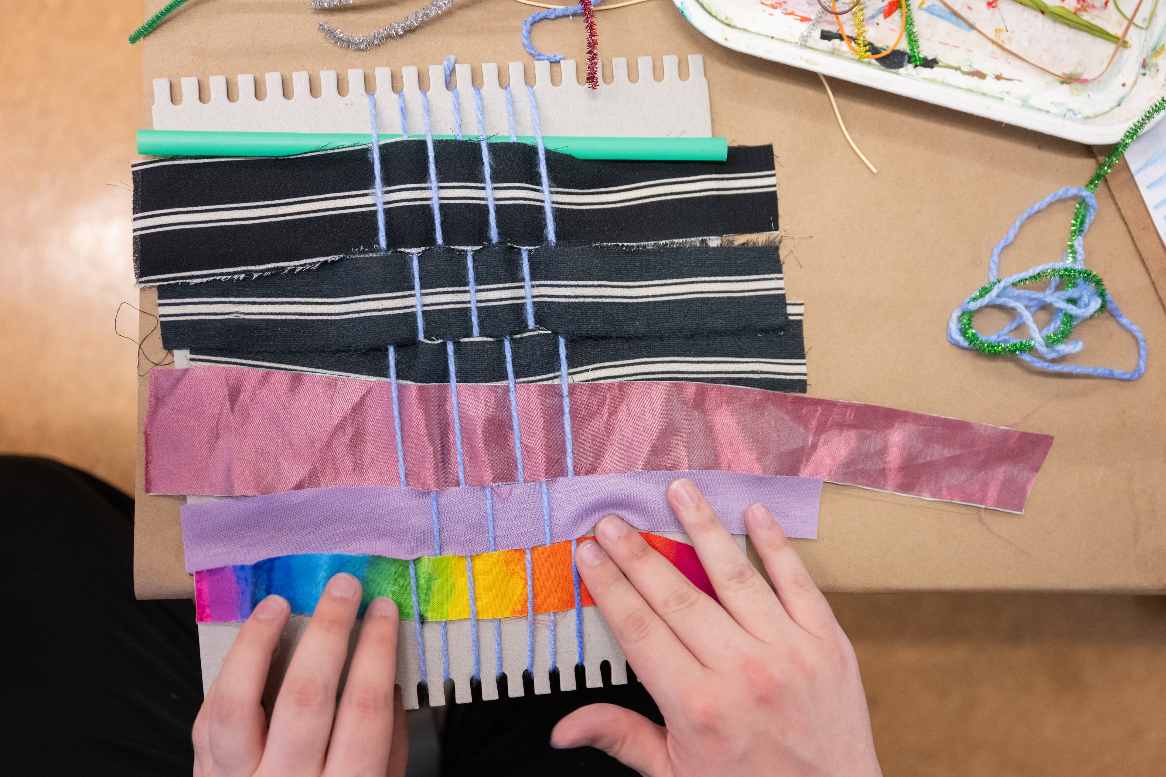 A warped weaving loom sits on an artmaking table; there are woven bits of black and white striped fabric, metallic pink fabric, light purple fabric, and an ombre rainbow fabric. The bottom of the loom, near the rainbow fabric, is held by two hands.