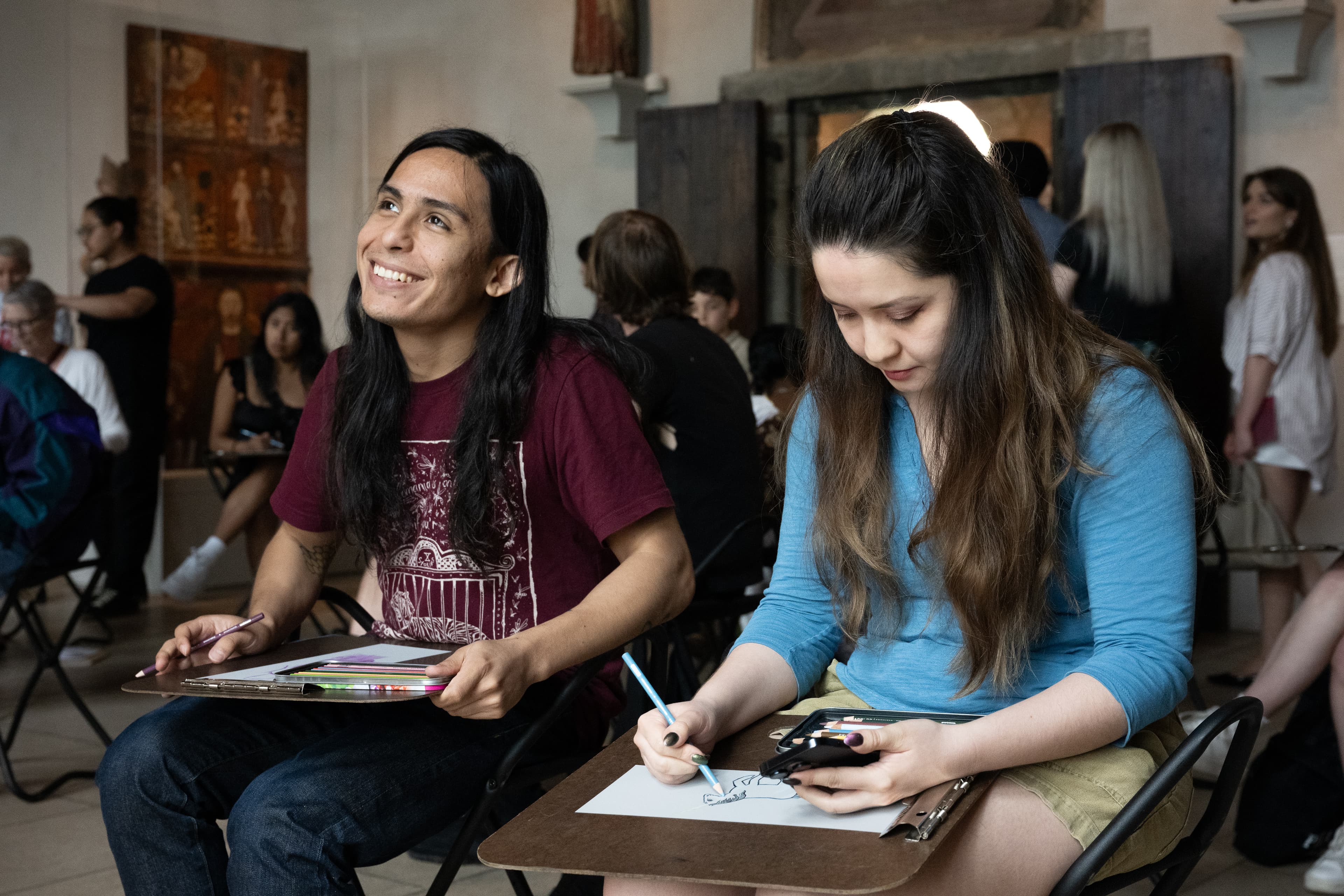Two people sitting in a gallery. One is wearing a dark red shirt and smiling, and the other is wearing a blue shirt and drawing.