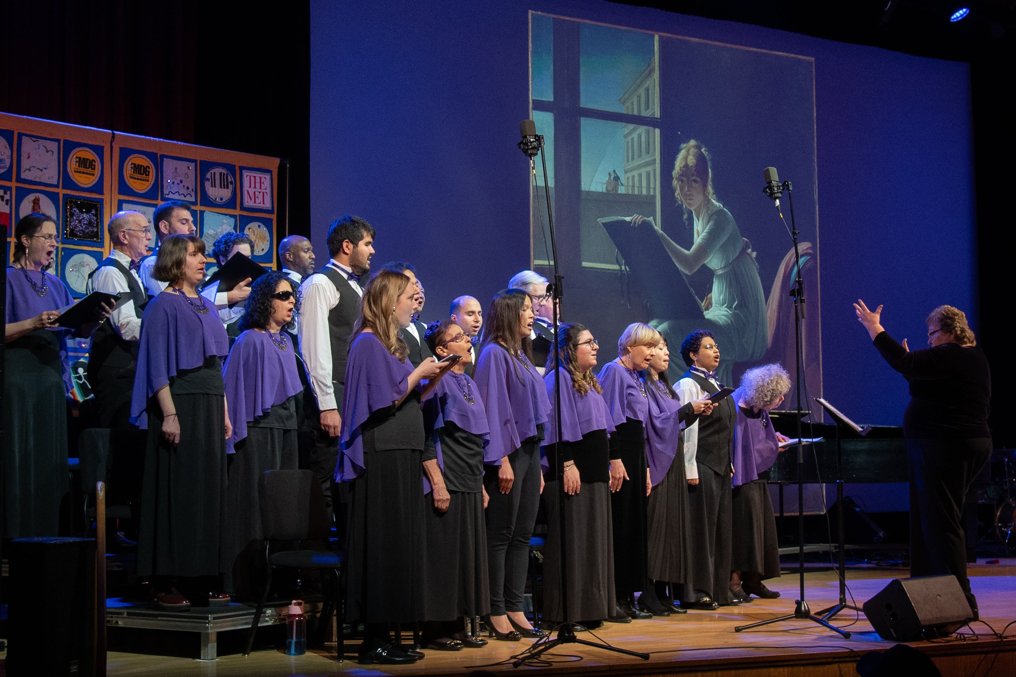 A group of performers dressed in purple and black stand on risers on a stage at an angle while a choir instructor stands in front of them to direct them. In the background, a portrait painting is projected on a screen behind them.