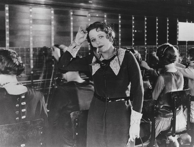 A black and white film still of a woman looking to the side, hand on her head, with a telephone receiver attached to her neck. A row of women managing telephone operator connection boards work behind her.