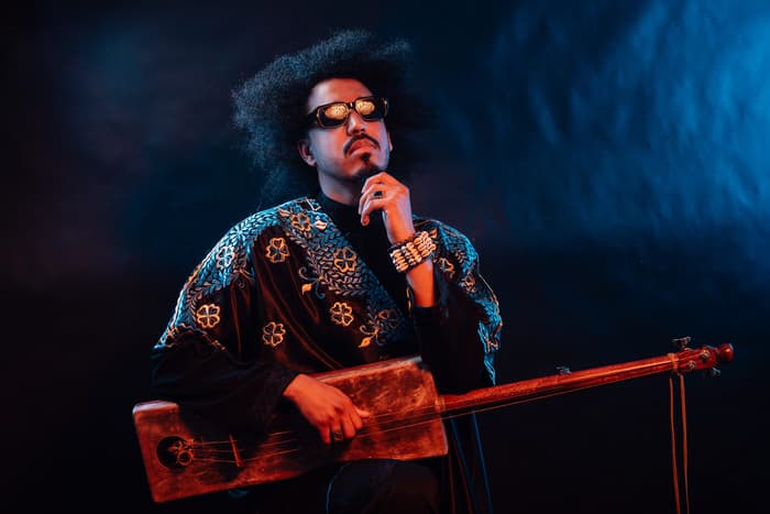 A man faces the camera holding his instrument, the gimbri or sinter, wearing a blue and black robe, bracelet and sunglasses.