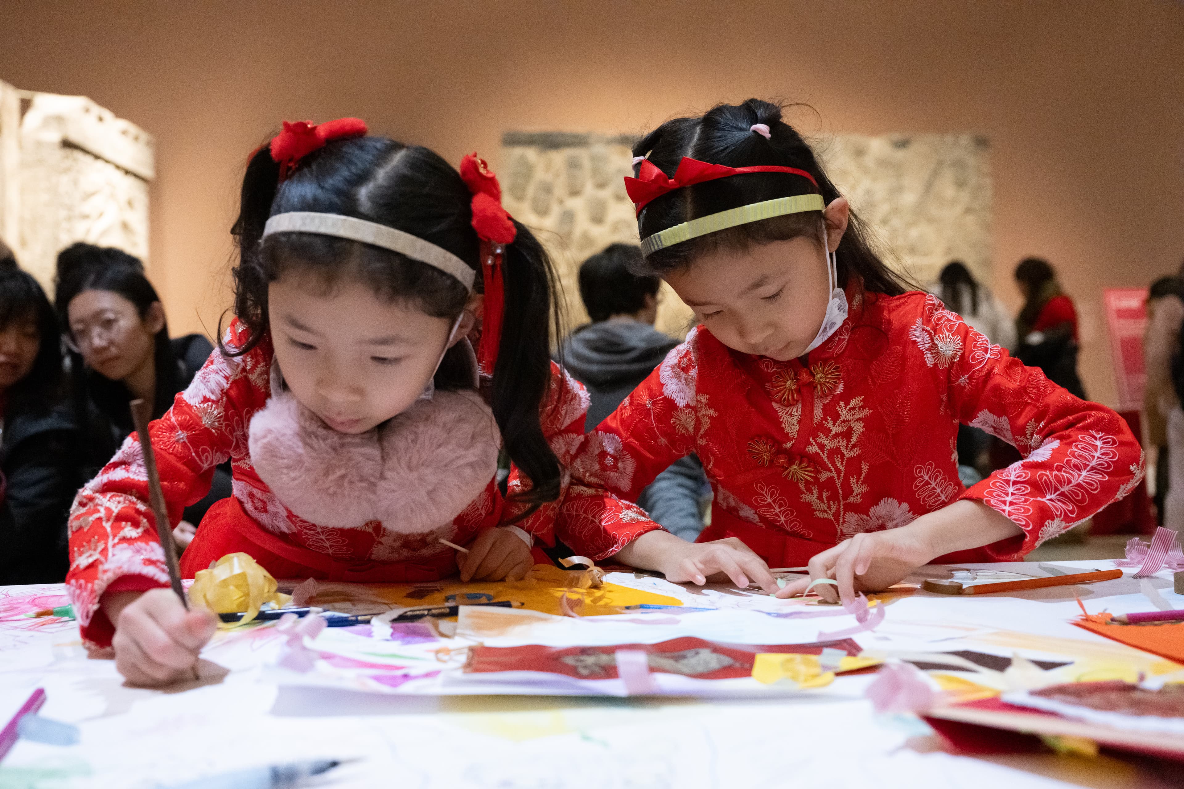 Two little girls wearing red clothing and red hair accessories work with intense focus on a paper craft activity for a past Lunar New Year Festival in one of the Met’s Asian Art galleries. Other festival attendees mill around in the background.