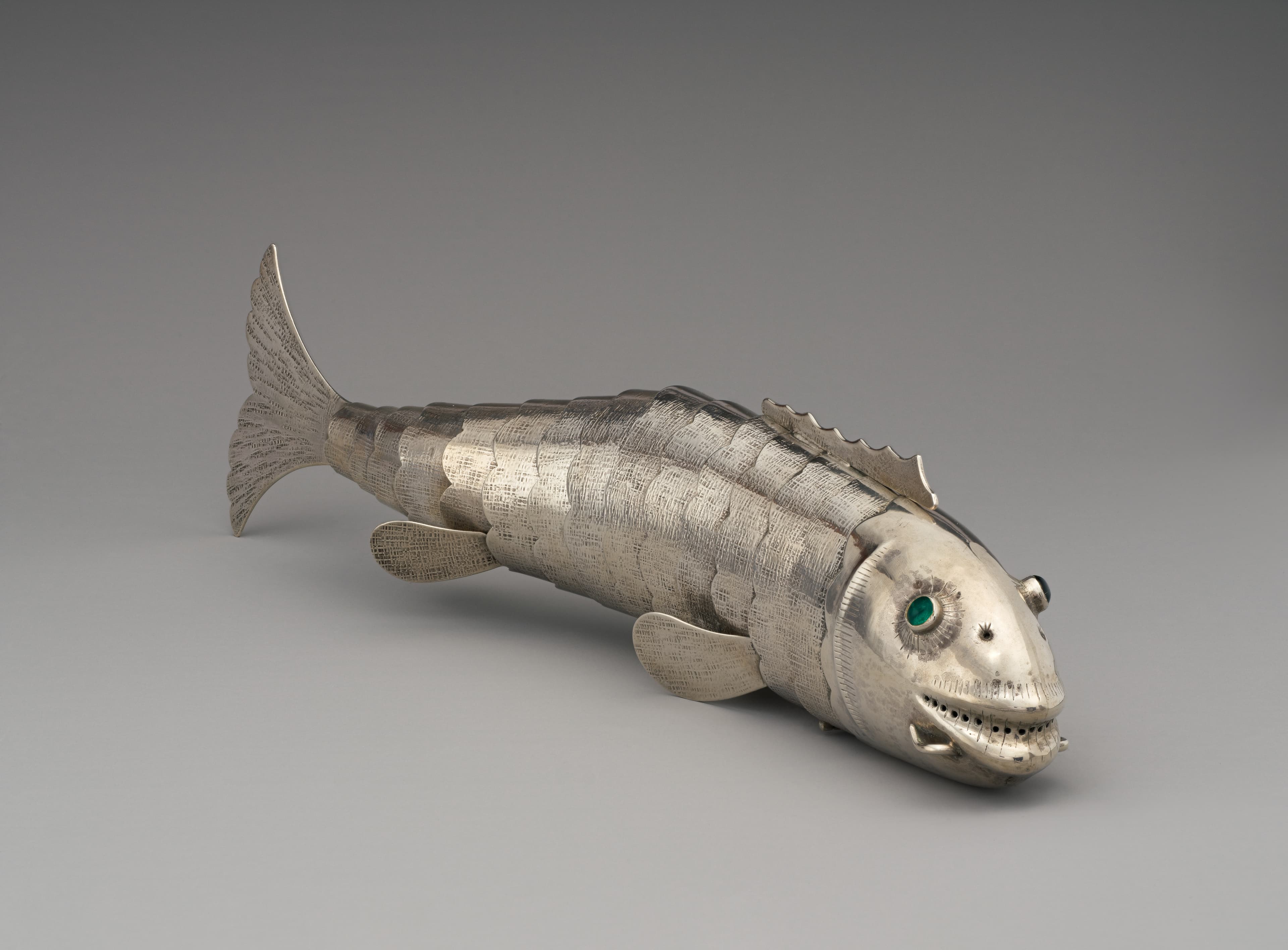 A silver fish-shaped vessel pointed at an angle towards the camera. The fish has emerald eyes and a smile.