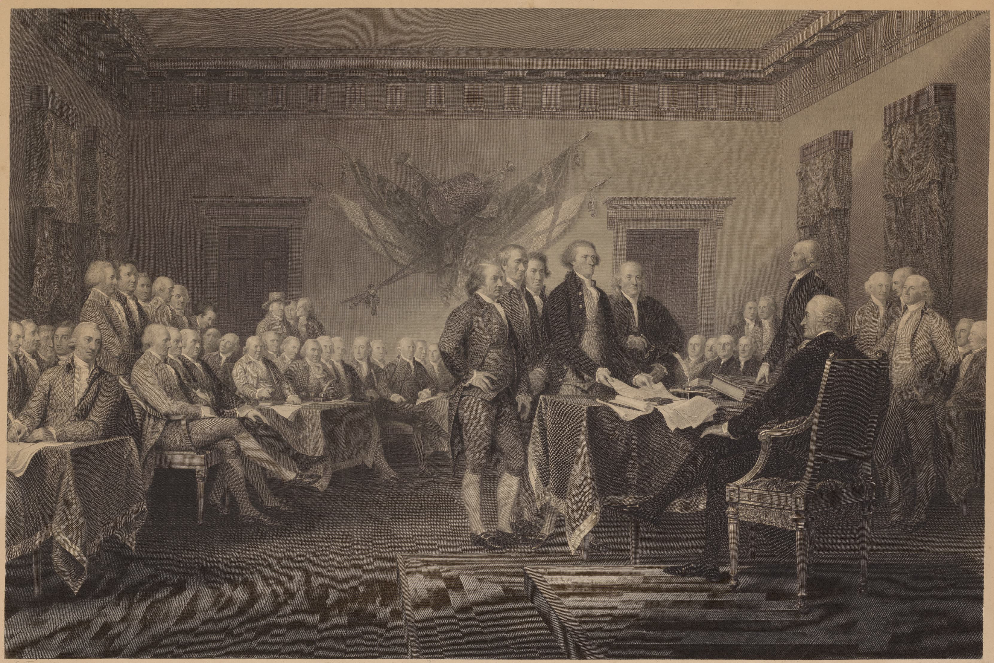 A print of the signing of the Declaration of Independence. Several founding fathers are visible standing around a table with papers on it, while more men sit in the background