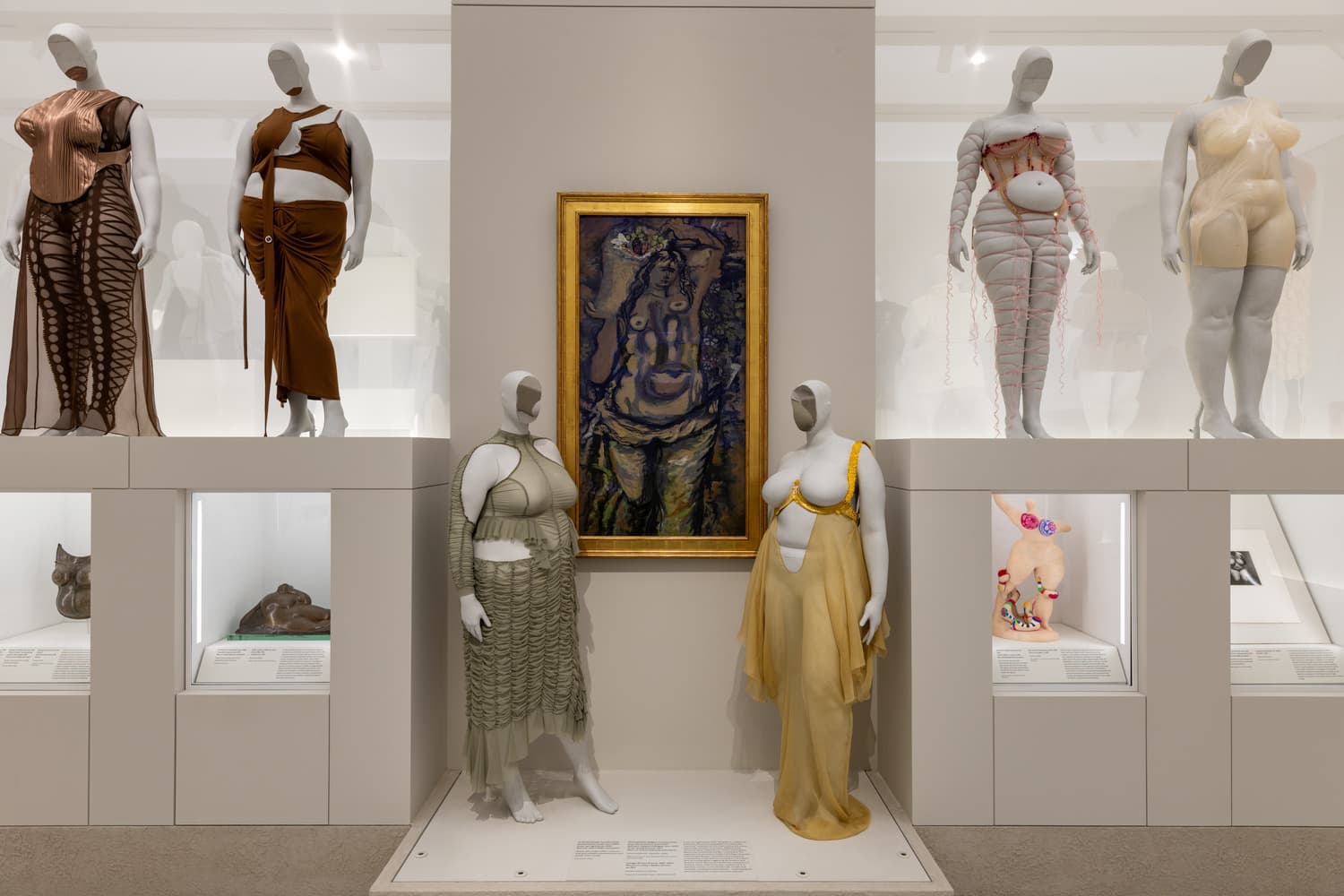 A tiered gallery installation featuring six mannequins of fat female bodies wearing ensembles, and five works of art in various media.